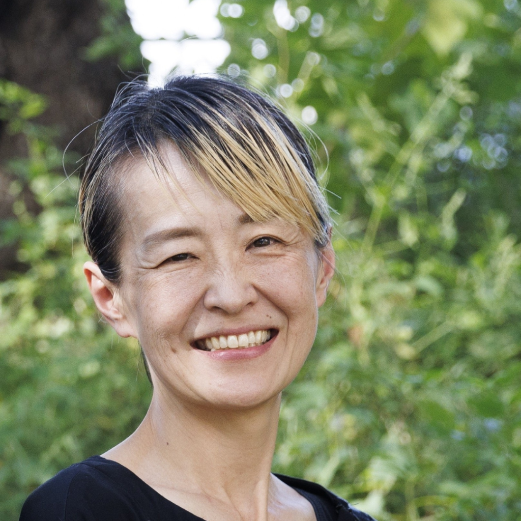 駒井由理子さんの顔写真。短い金髪のハイライトが入った髪型の女性で、緑の自然を背景に微笑んでいる。