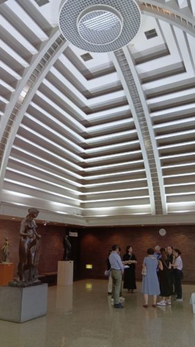美術館エントランスでグループが輪になって話している写真。画面にはエントランスの高くて白いドーム型の天井が上まで写されている。