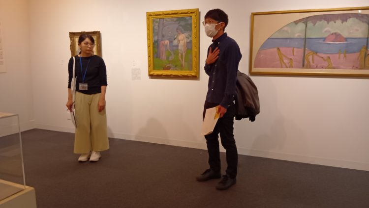 事前の打ち合わせで手話通訳者の立ち位置を確認している様子の写真。作品を挟むように二人の人物が立っている。左側は学芸員の女性、右側は手話通訳の男性。