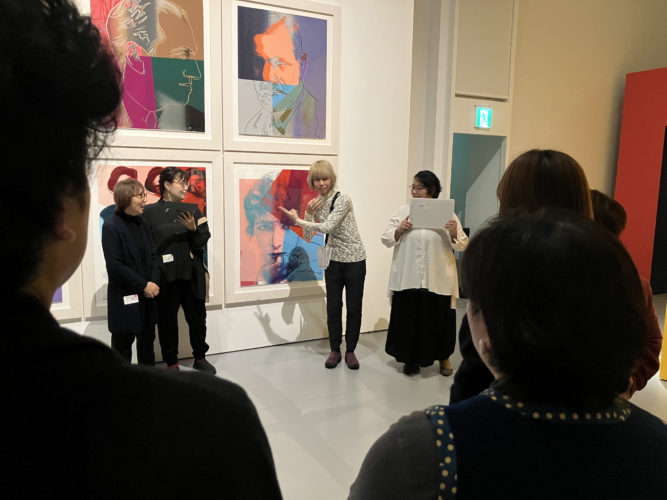 会場となった展示室で、色彩豊かな人物肖像作品の前にファシリテーター２名と手話通訳者２名が立ち、参加者に向けて手話で自己紹介をしている様子の写真。