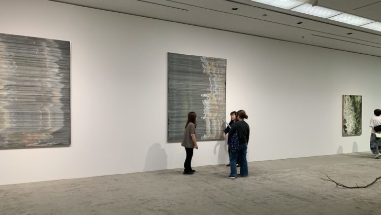 「Nerhol」展にて、手話通訳者同士が手話表現の最終確認をしている様子の写真。白い壁に展示された大きな作品の前に、手話通訳者2名とみんミスタッフが立って話している。