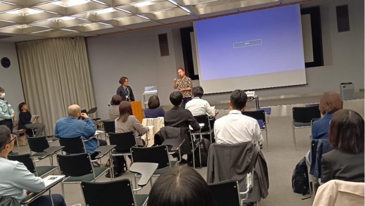 スクリーンを背に登壇者が手話で話している様子の写真。研修参加者は並べられた椅子に座っている。