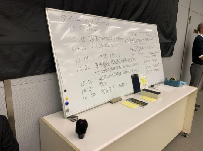 会場の後方の机にホワイトボードが机に立てかけてある写真。タイムスケジュールが手書きで書かれている。