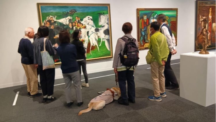 絵画の前を研修参加者が集まって対話しながら鑑賞をしている様子の写真。当事者の協力者のなかには盲導犬を連れた人が参加している。