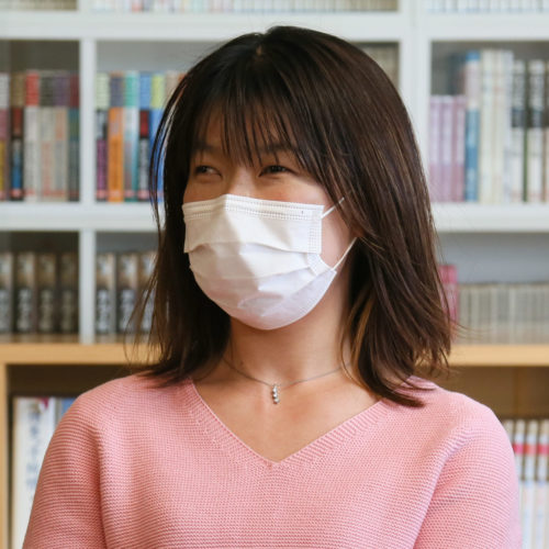 書棚を背景に、ピンク色のセーターを着た人物がマスクを着用して着席し、話をしている様子の写真