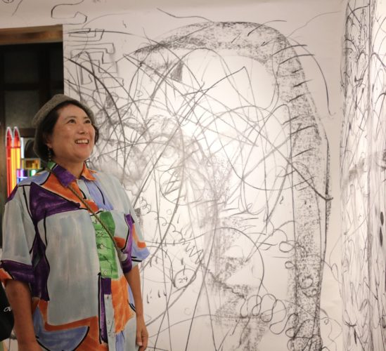 大規模な壁面ドローイング作品の前で、カラフルなシャツを着た人物が作品を見上げている写真