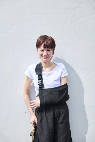 白い壁を背景に、笑顔で立つ平田春果さんのポートレート写真。赤茶色のショートヘアで、白い半袖シャツと黒いパンツを着用し、左腕を黒いアームスリングで支え、右手に杖を持っている。
