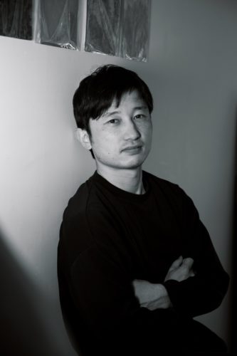 黒い長袖の服を着た飯田竜太氏のモノクロのポートレート写真。壁を背に、腕を組んでカメラの方向を見ている。
