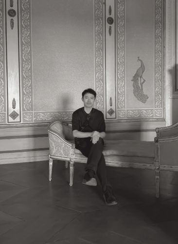 磯谷博史氏のポートレート写真。装飾的な壁面のある室内で、黒い服を着た磯村氏が脚を組み、正面を向いて座っているモノクロ写真。壁面には植物模様の縁取りや孔雀の絵が描かれている。